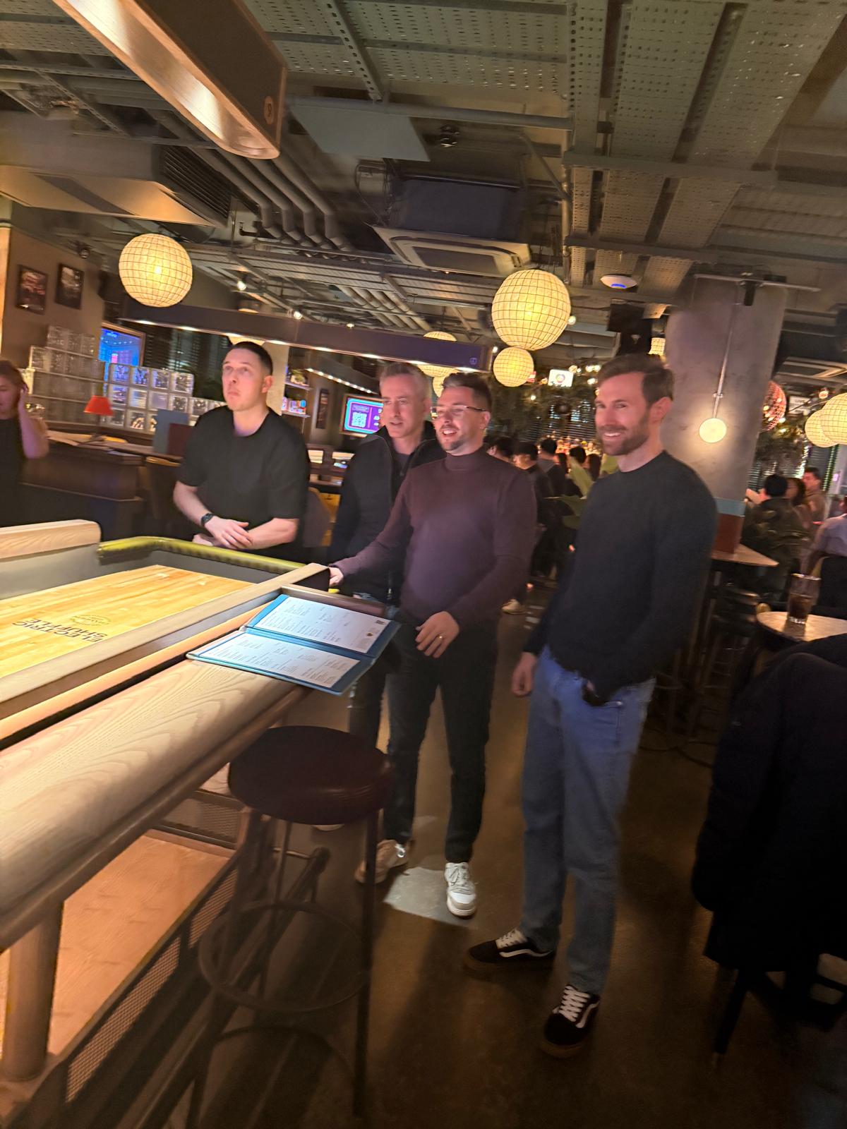 Four members around the shuffleboard table, waiting on the next puck
