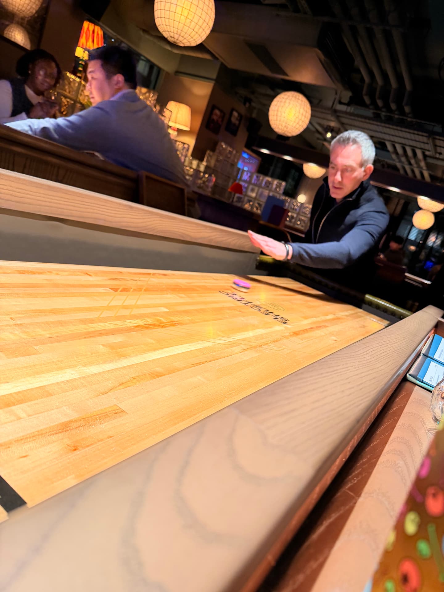 A member sliding a puck down the shuffleboard table while others talk at the bar